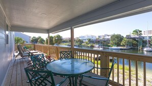 A covered outdoor deck with a table and chairs, overlooking a marina with boats and buildings in the background. - The Back Porch (Destin)