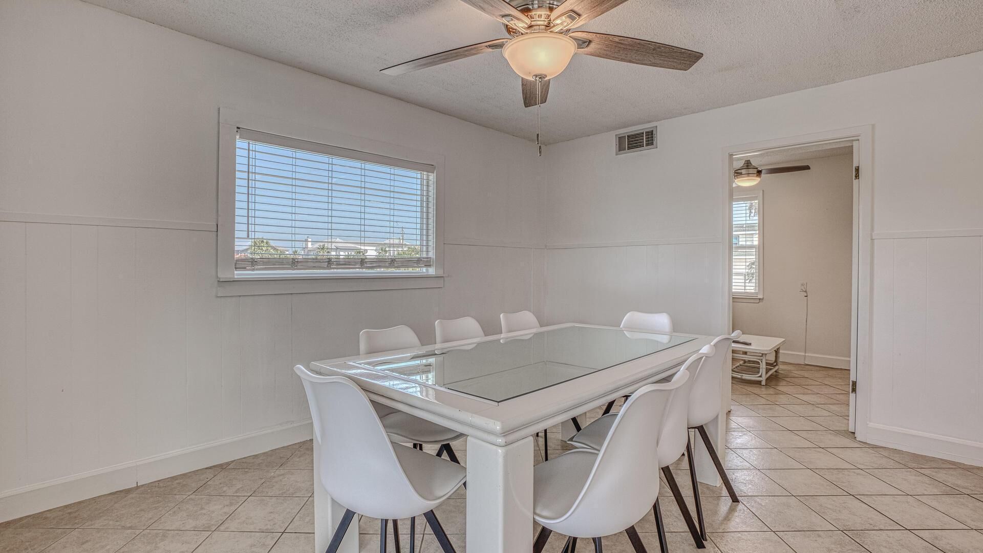 A spacious room with a large glass-top table surrounded by white chairs, a ceiling fan, and a window with blinds.