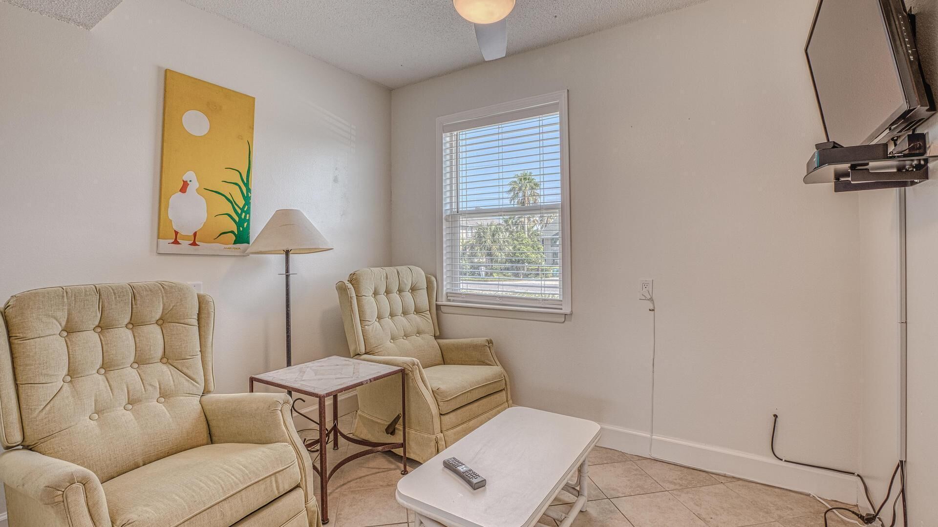 A living room with two beige armchairs, a small table, and a window with blinds.