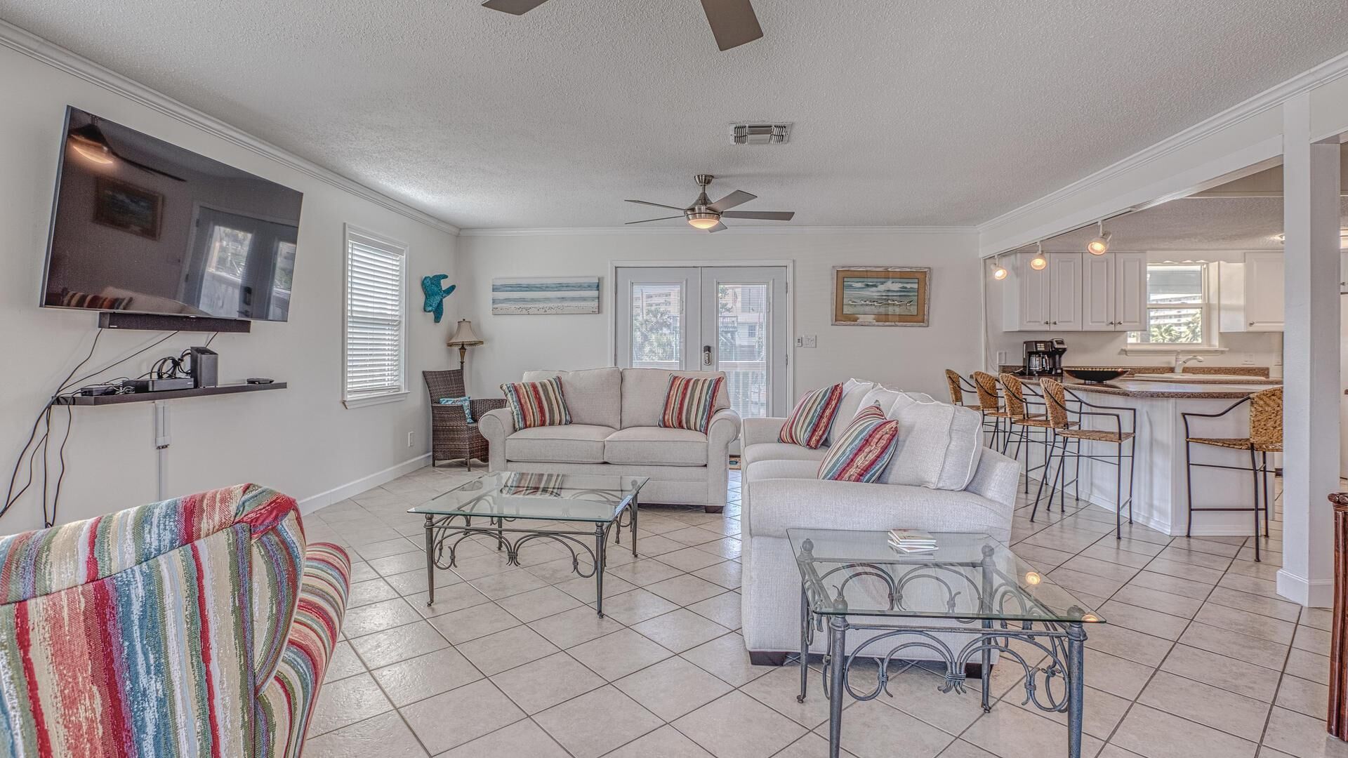 A spacious living room with a large sofa, a coffee table, and a television mounted on the wall. There is a dining area with a kitchen in the background.