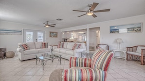 A spacious living room with a sofa, armchairs, a coffee table, and a dining area. There are ceiling fans, a kitchen in the background, and a framed picture on the wall. - The Back Porch (Destin)