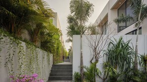 Een smal pad dat leidt naar een gebouw met een modern ontwerp, omzoomd door weelderige begroeiing en een bloembed met roze bloemen.
