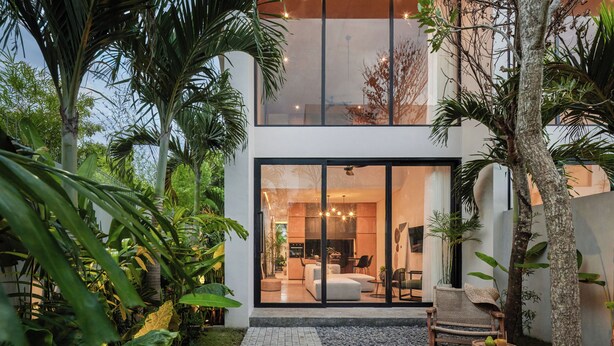Een modern huis met een glazen gevel, een terras met zitplaatsen en weelderige begroeiing.