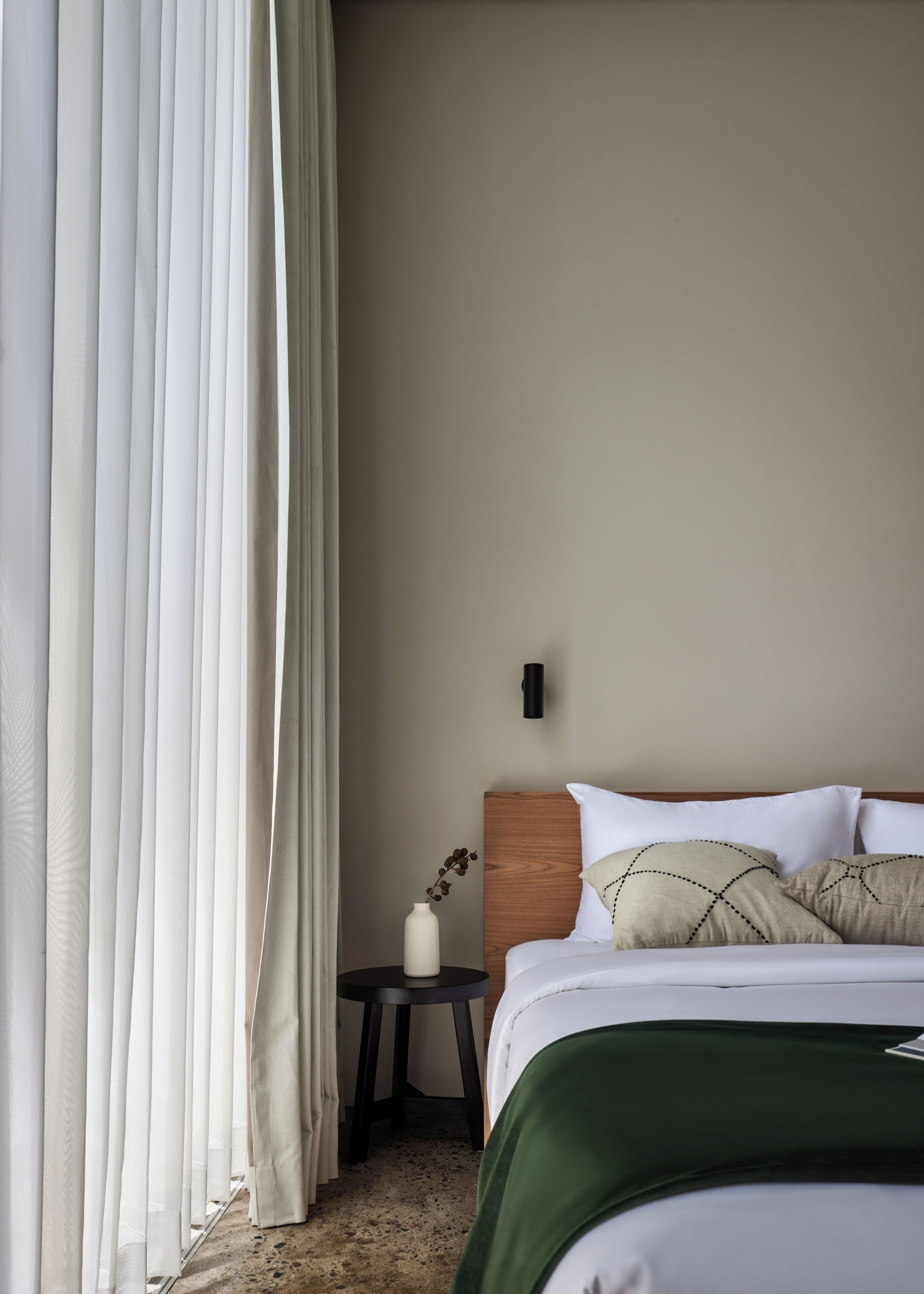 Quarto de hotel com uma cama, uma mesa de cabeceira, um vaso com planta e cortinas translúcidas.