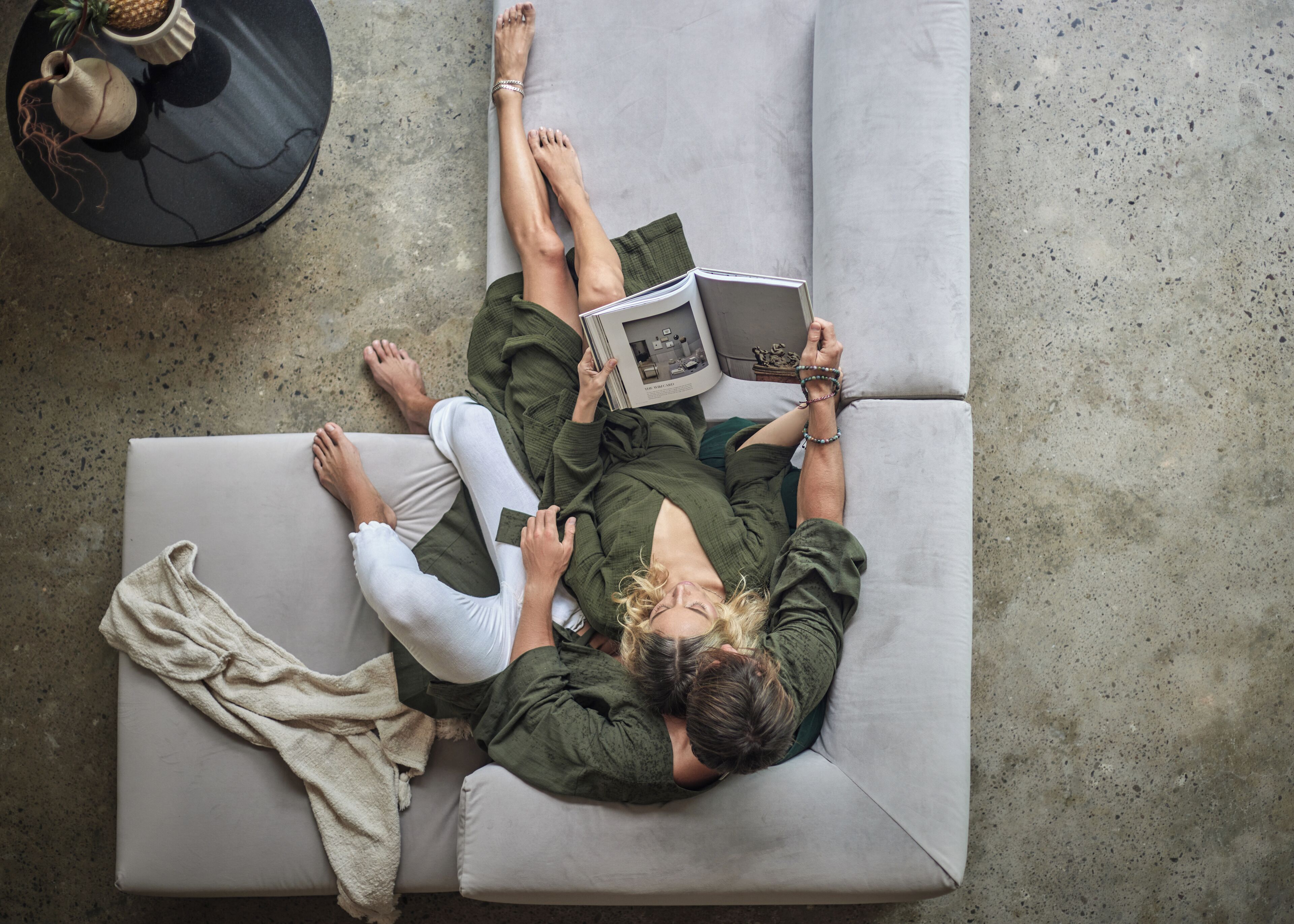 A person lying on a grey sofa with a book, wearing a green top and white pants, with a small table and a plant in the background.