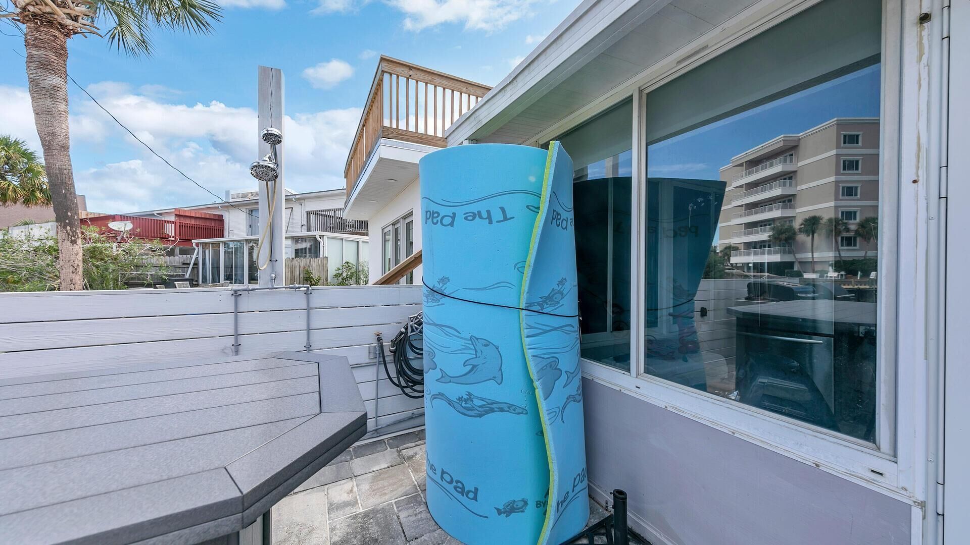 A blue surfboard leaning against a building with a window, a wooden deck, and a palm tree in the background.