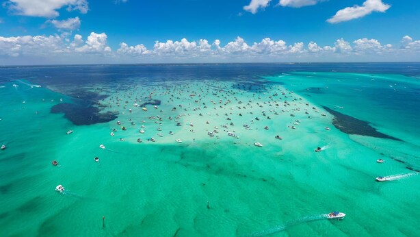 Vista aérea de una laguna turquesa con numerosas embarcaciones, rodeada de aguas azules cristalinas y algunas nubes dispersas.