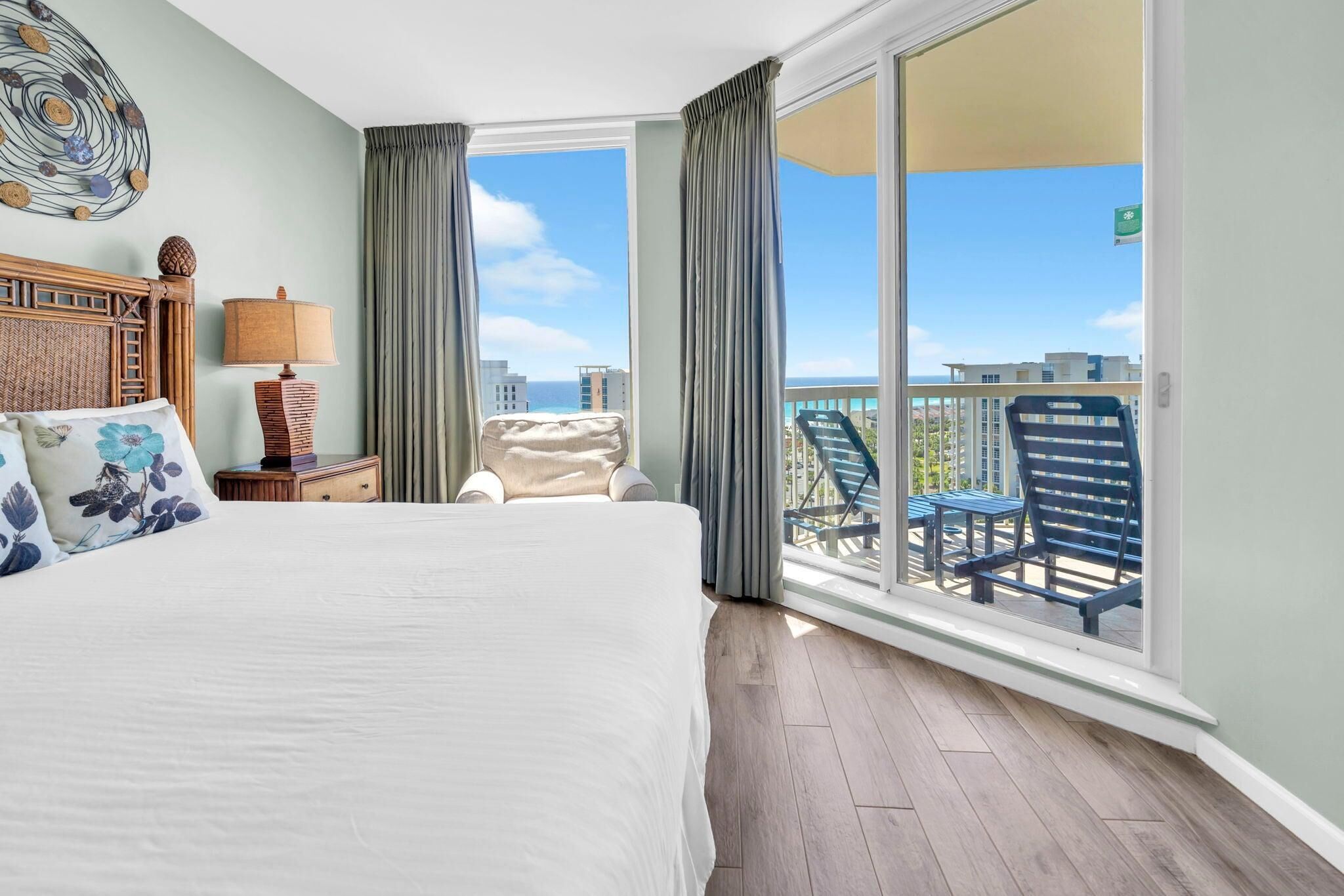 A hotel room with a bed, a nightstand, a lamp, a chair, and a balcony with a view of buildings and the sea.