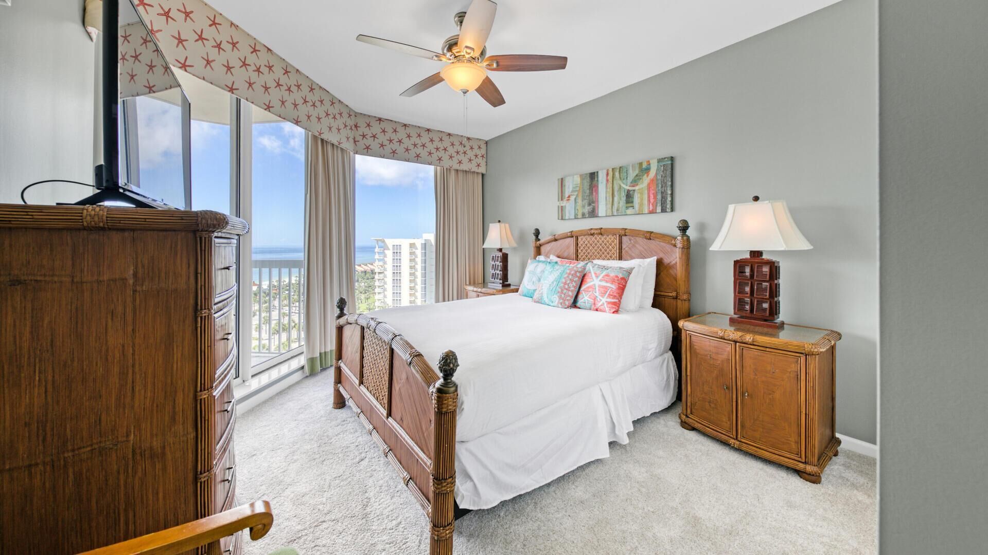 A bedroom with a large bed, a wooden wardrobe, a nightstand, a ceiling fan, and a view of the city.