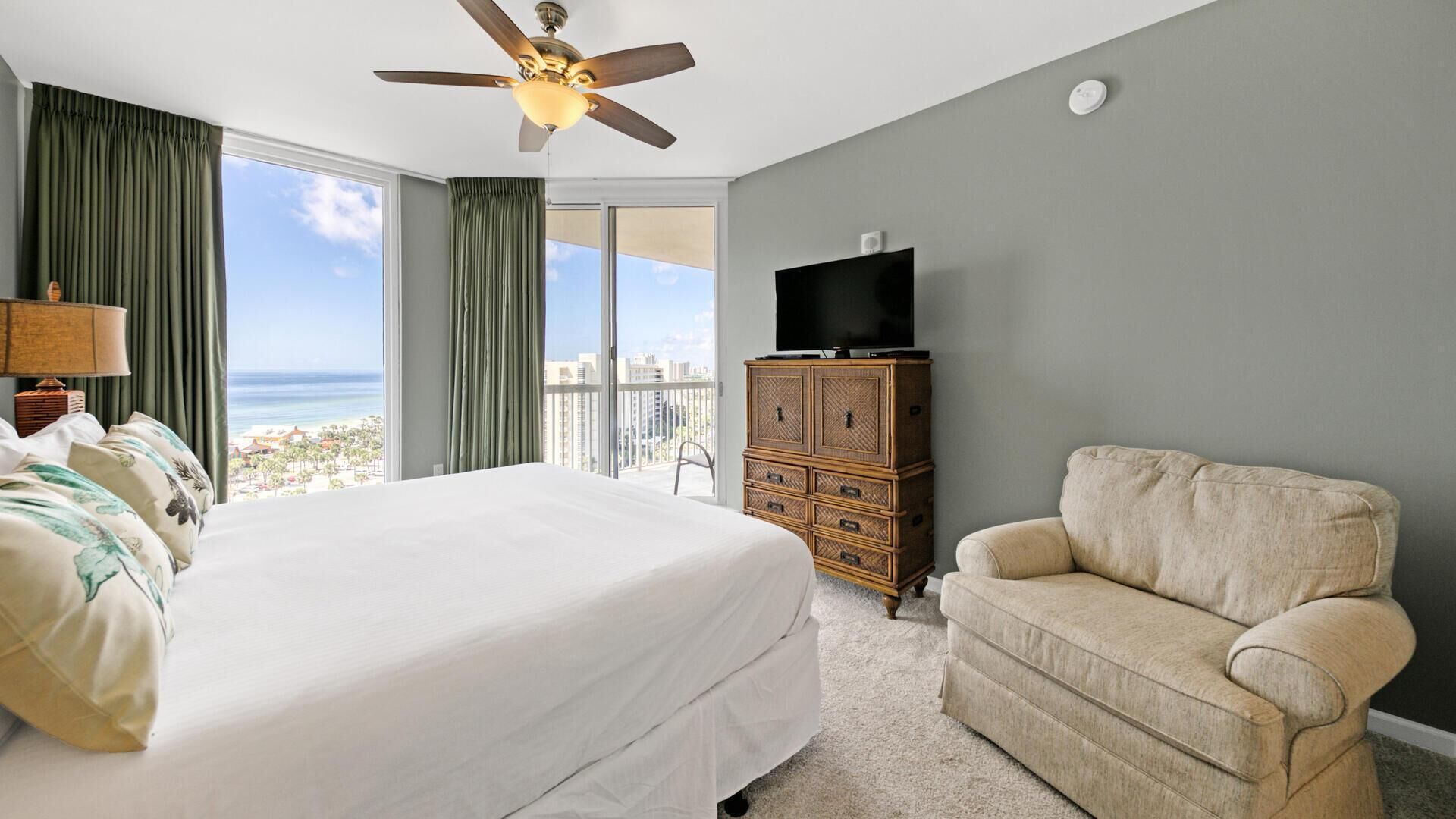 Une chambre d’hôtel avec un grand lit, un ventilateur de plafond, un téléviseur posé sur une commode en bois et une vue sur l’océan.