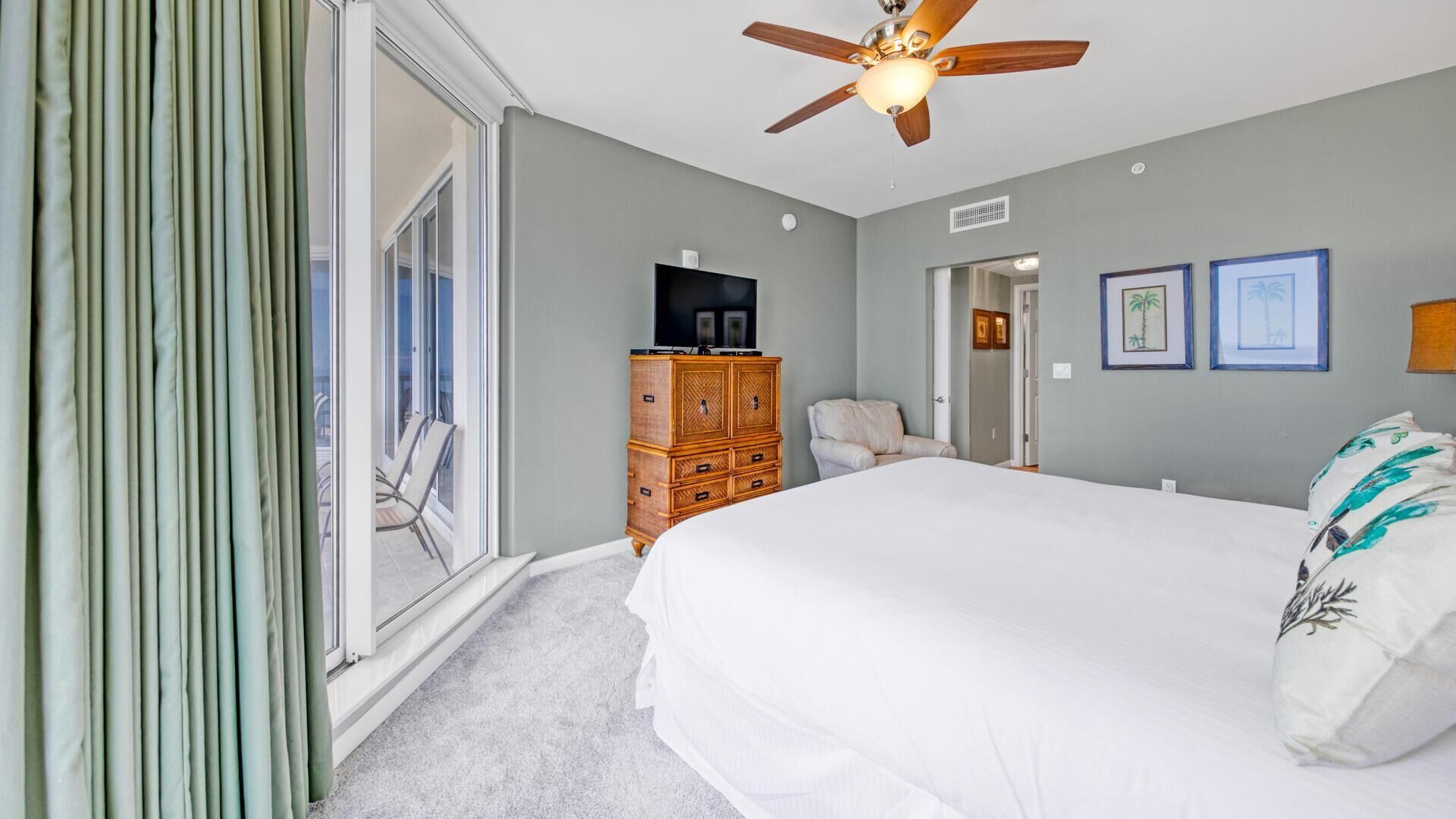 A bedroom with a large bed, a wooden dresser, a ceiling fan, and a view through a sliding door to an outdoor area.