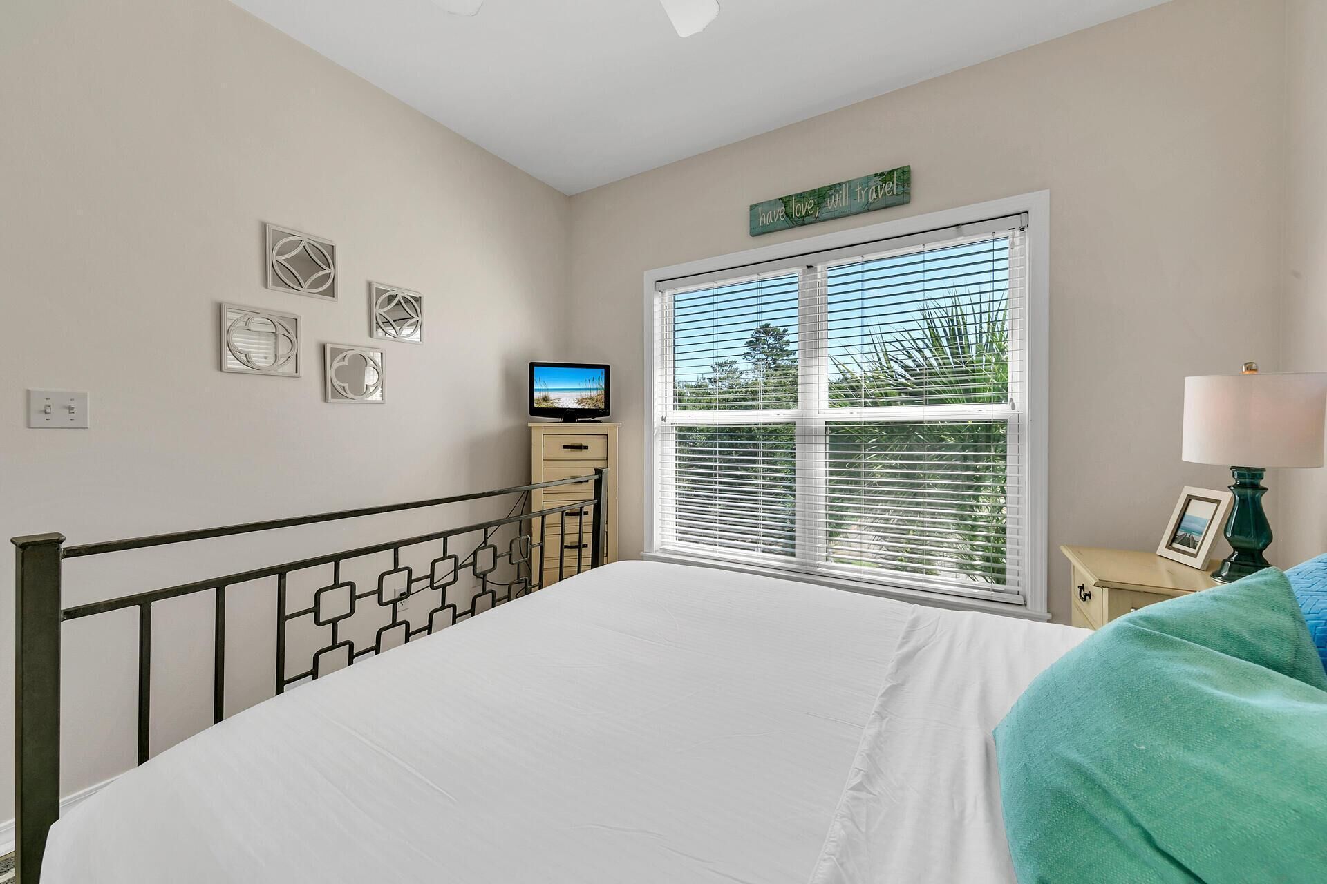 A bedroom with a bed, a TV, a window with blinds, a bedside table with a lamp, and wall decorations.