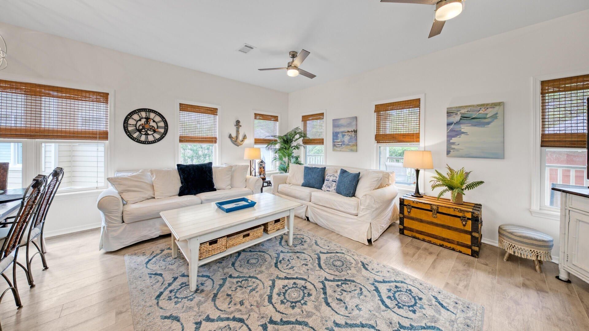 A spacious living room with two white sofas, a coffee table, and a wooden chest of drawers. There are large windows with blinds, a ceiling fan, and a clock on the wall.