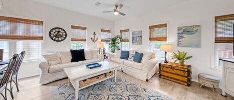 A spacious living room with two white sofas, a coffee table, and a wooden chest of drawers. There are large windows with blinds, a ceiling fan, and a clock on the wall.