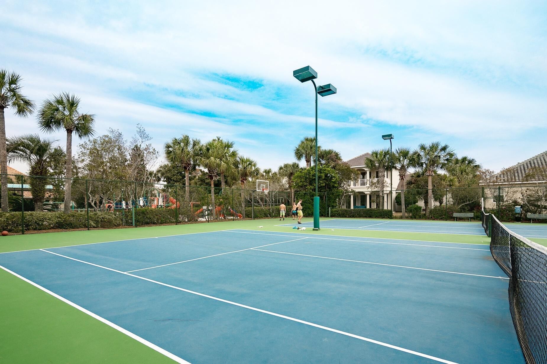 An outdoor tennis court with a green and blue surface, surrounded by palm trees and a fence.