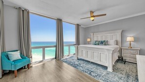 Un dormitorio con un amplio ventanal con vistas al océano, una cama con cabecera blanca, un sillón azul turquesa y una mesita de noche con lámpara.
