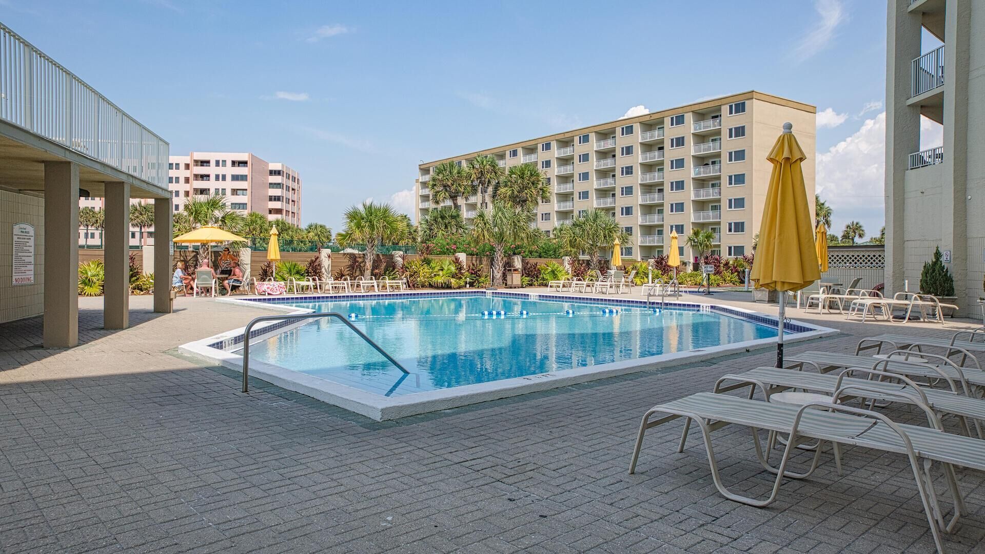 A swimming pool with lounge chairs and umbrellas, surrounded by buildings and greenery.