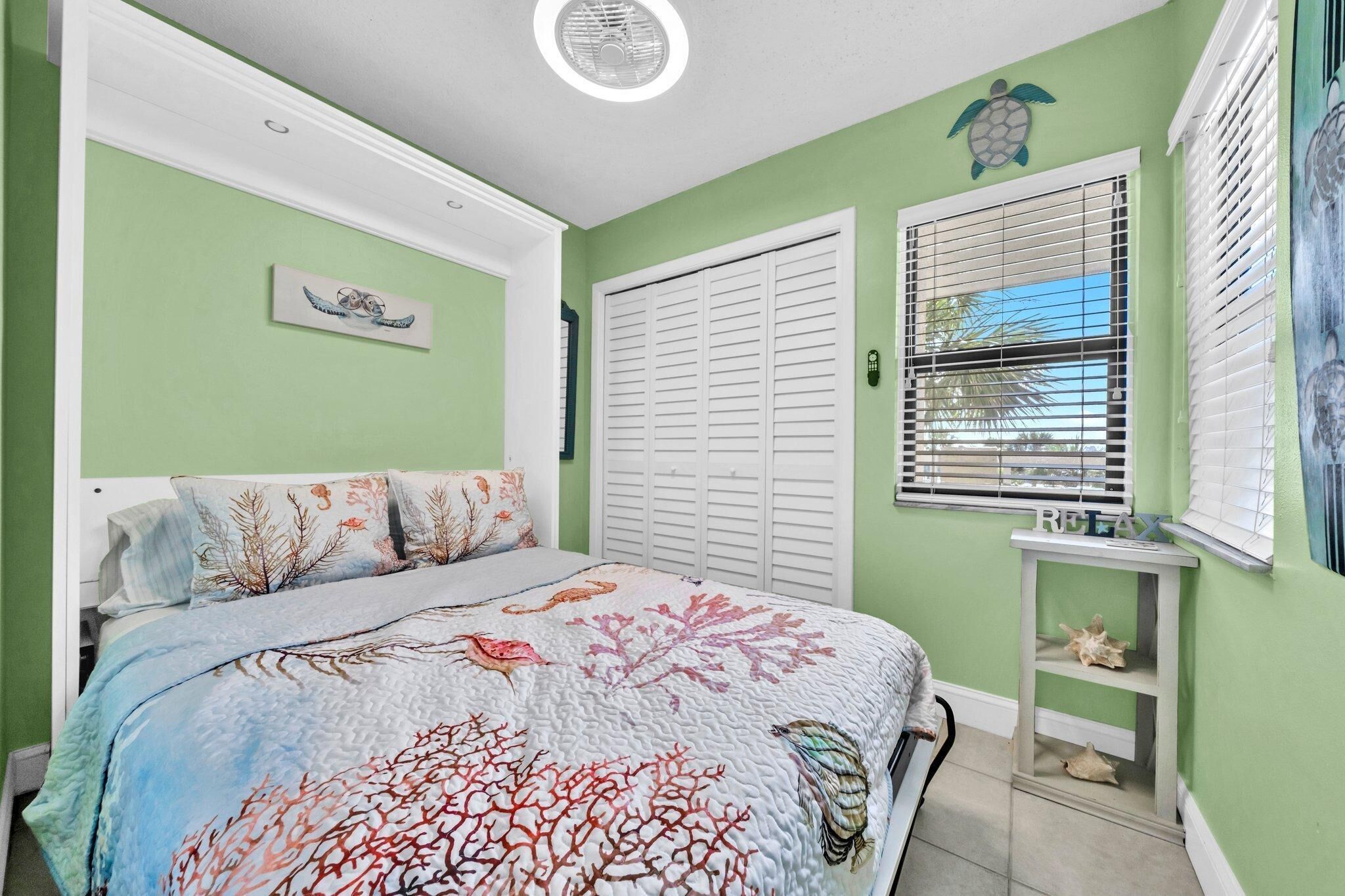 A bedroom with a bed, a window with blinds, a small table, and a wall decoration.