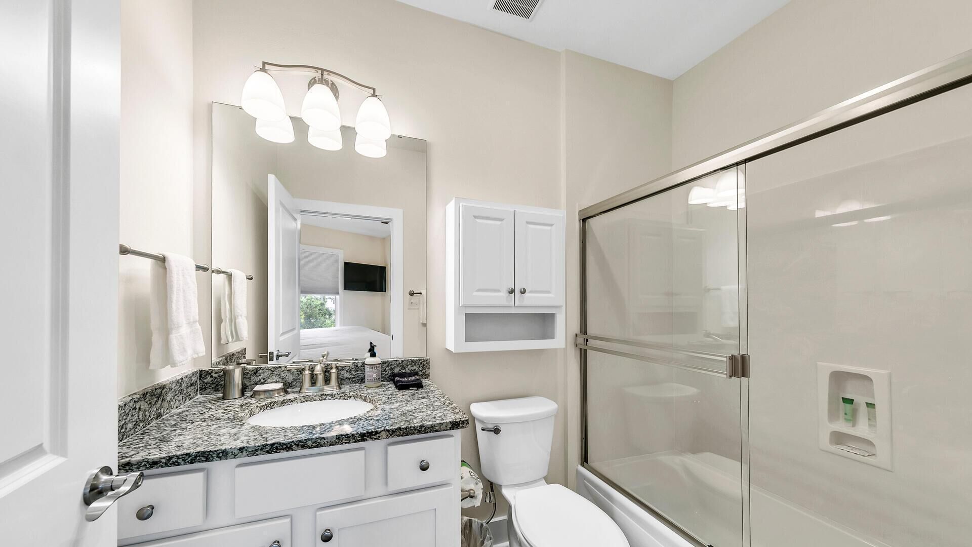 Un baño moderno con lavamanos de granito, inodoro, cabina de ducha con mampara de vidrio y tocador con espejo e iluminación superior.