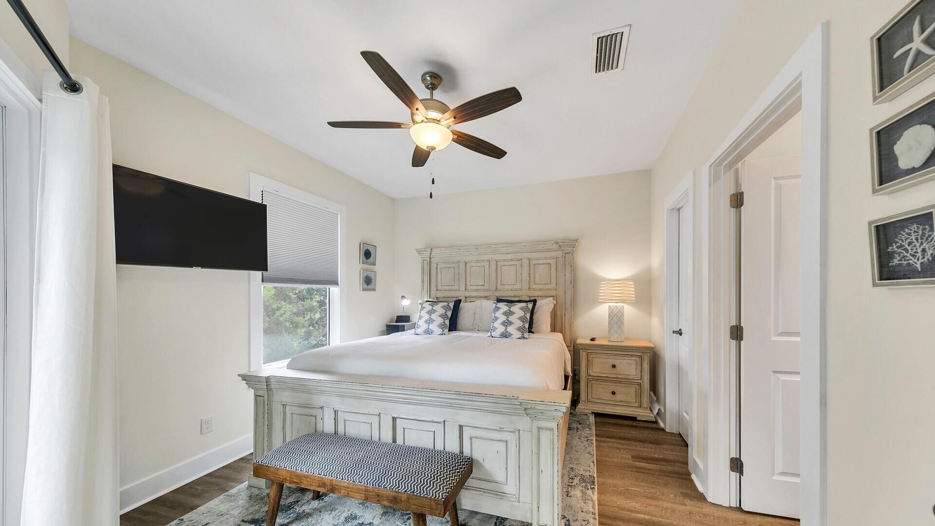 A bedroom with a bed, a bench, a ceiling fan, a TV, and a window.