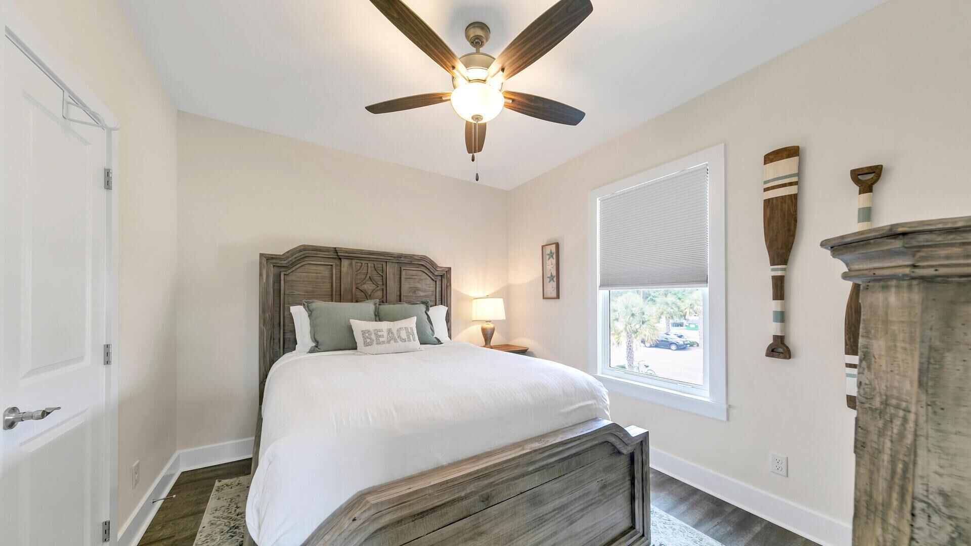 Une chambre à coucher avec une tête de lit en bois, un lit avec une literie blanche, un ventilateur de plafond, une fenêtre avec des stores et une table de chevet avec une lampe.
