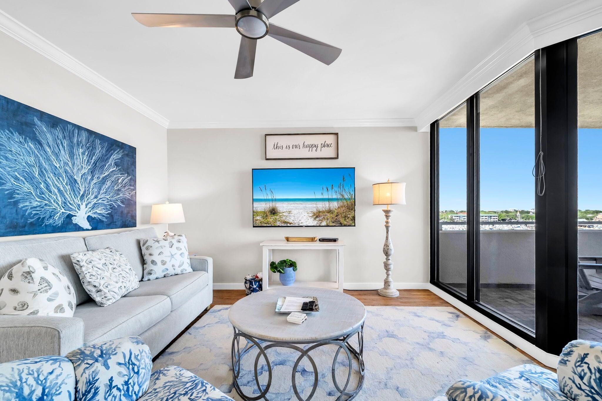 A modern living room with a sofa, a round coffee table, a ceiling fan, a TV, and a large window with a view.