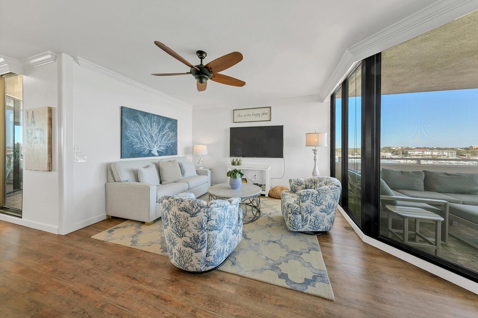A spacious living room with a large window, a ceiling fan, a white sofa, two patterned armchairs, a coffee table, and a painting on the wall.
