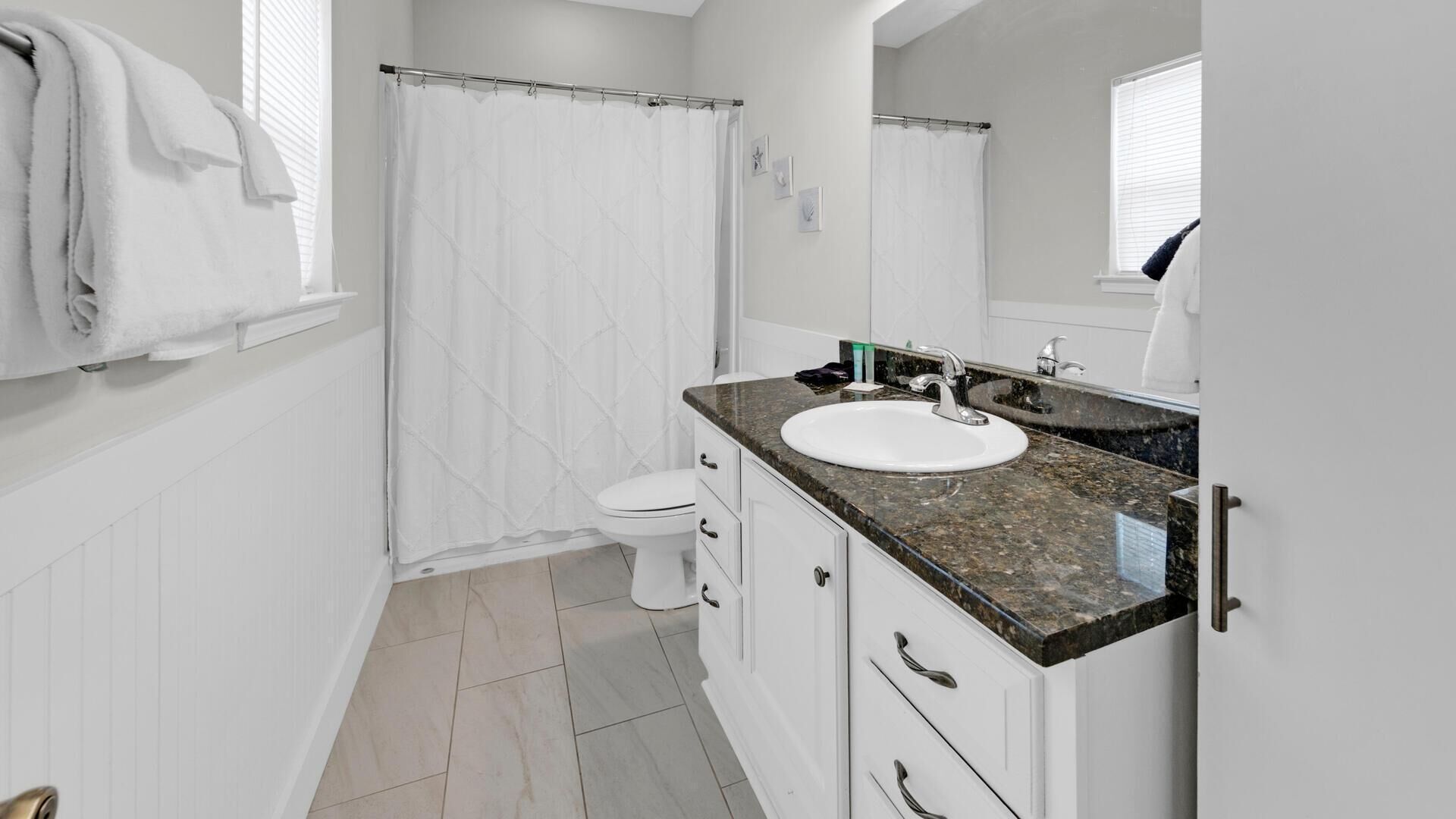 Une salle de bain moderne avec des toilettes blanches, une douche avec un rideau blanc, un lavabo sur un plan de travail en granit et des meubles blancs.