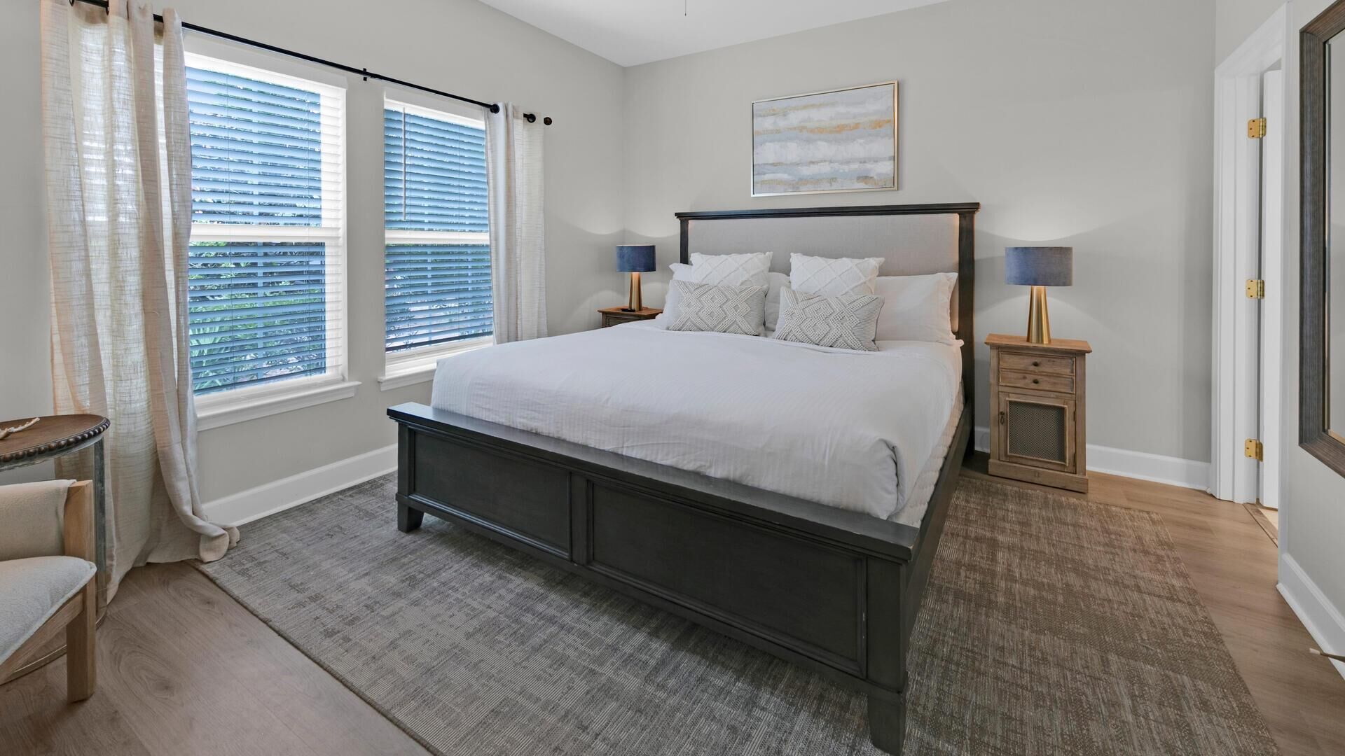 A bedroom with a large bed, two bedside tables, a chair, and a window with blinds.