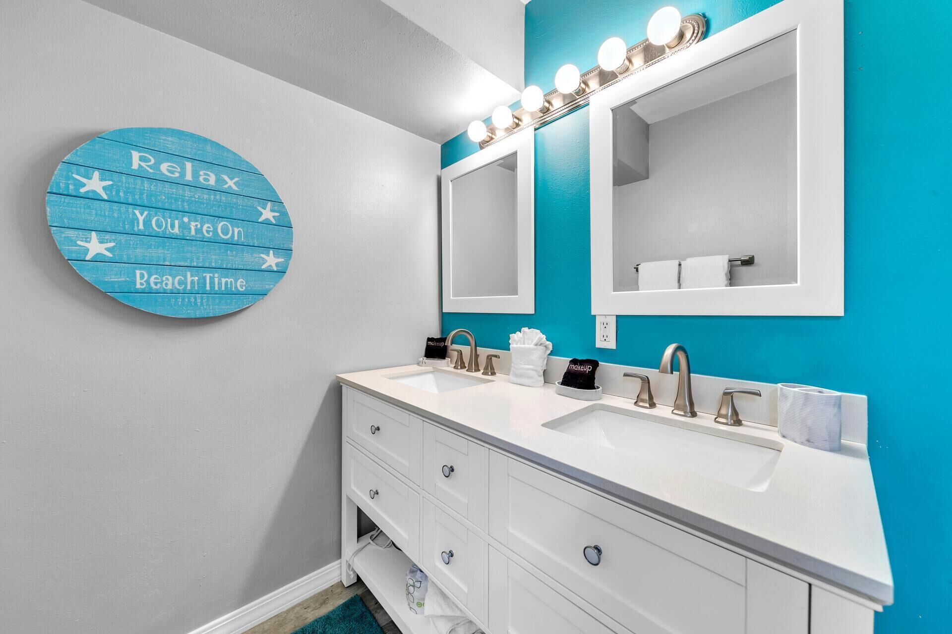 A bathroom with a vanity, two sinks, and a mirror with lights. There is a circular sign on the wall with text.