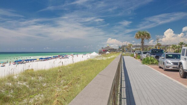 A beach with a paved walkway, beach umbrellas, and parked cars along the side. - Playa Blanca (Miramar Beach)