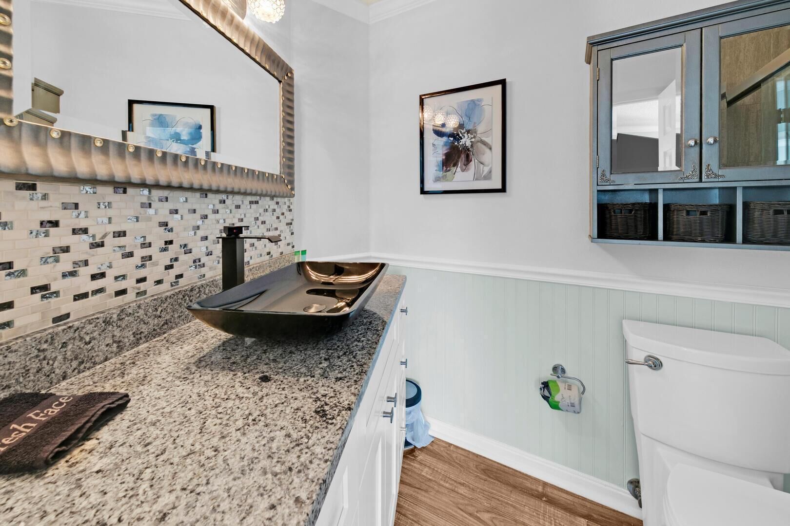 A bathroom with a granite countertop, a large mirror, a framed artwork, and a white toilet.