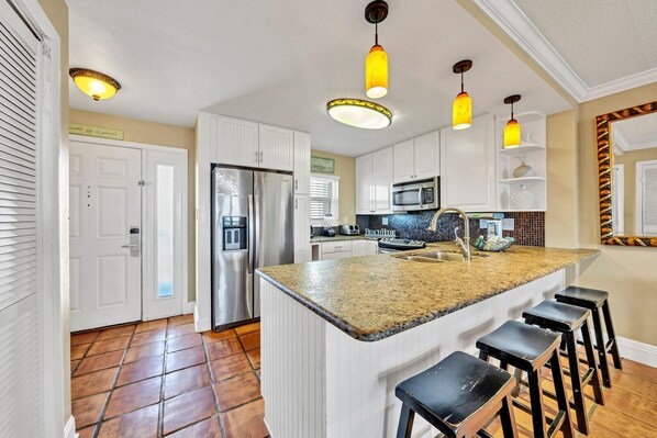 A modern kitchen with white cabinets, a stainless steel refrigerator, a built-in microwave, a granite countertop, and a bar stool area. - Emerald Isle 706 (Fort Walton Beach)