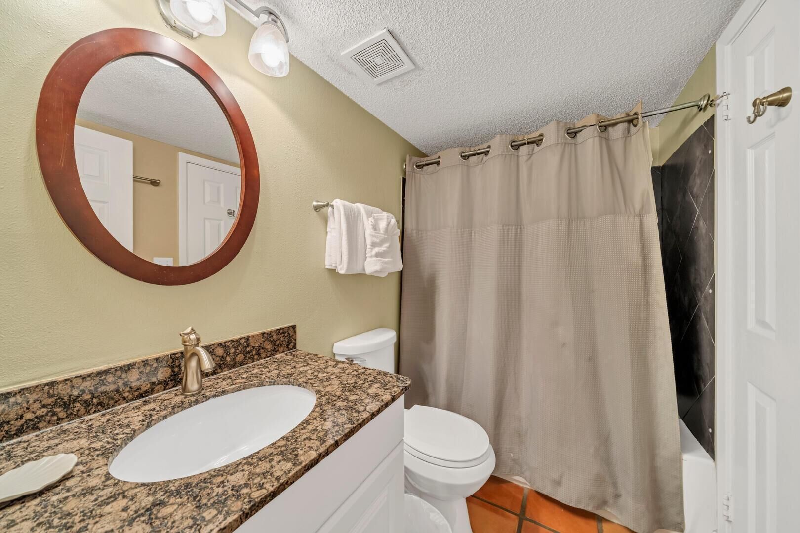 Un baño con lavamanos de granito, inodoro blanco, cortina de ducha color beige y espejo redondo.