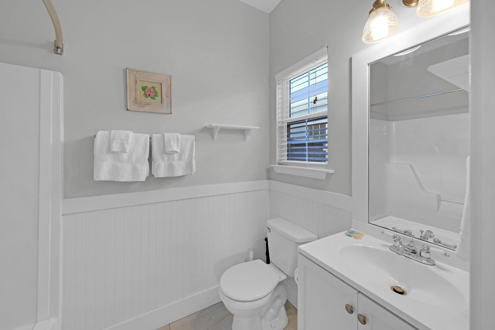 Ein Badezimmer mit Toilette, Waschbecken und Fenster mit Jalousien. Handtücher hängen an der Wand und ein Bilderrahmen ist zu sehen.