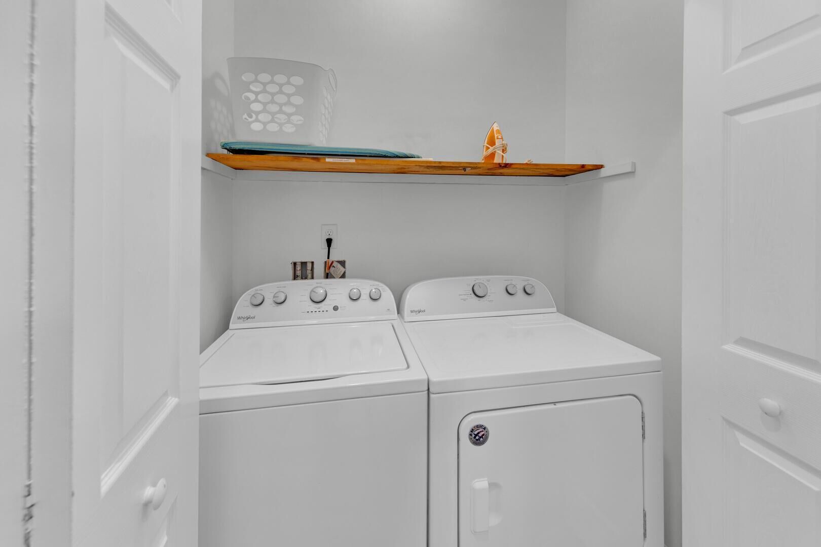 A laundry room with two white Whirlpool washers and dryers, a wooden shelf with a surfboard and a decorative item, and a power outlet on the wall.