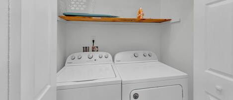 A laundry room with two white Whirlpool washers and dryers, a wooden shelf with a surfboard and a decorative item, and a power outlet on the wall.