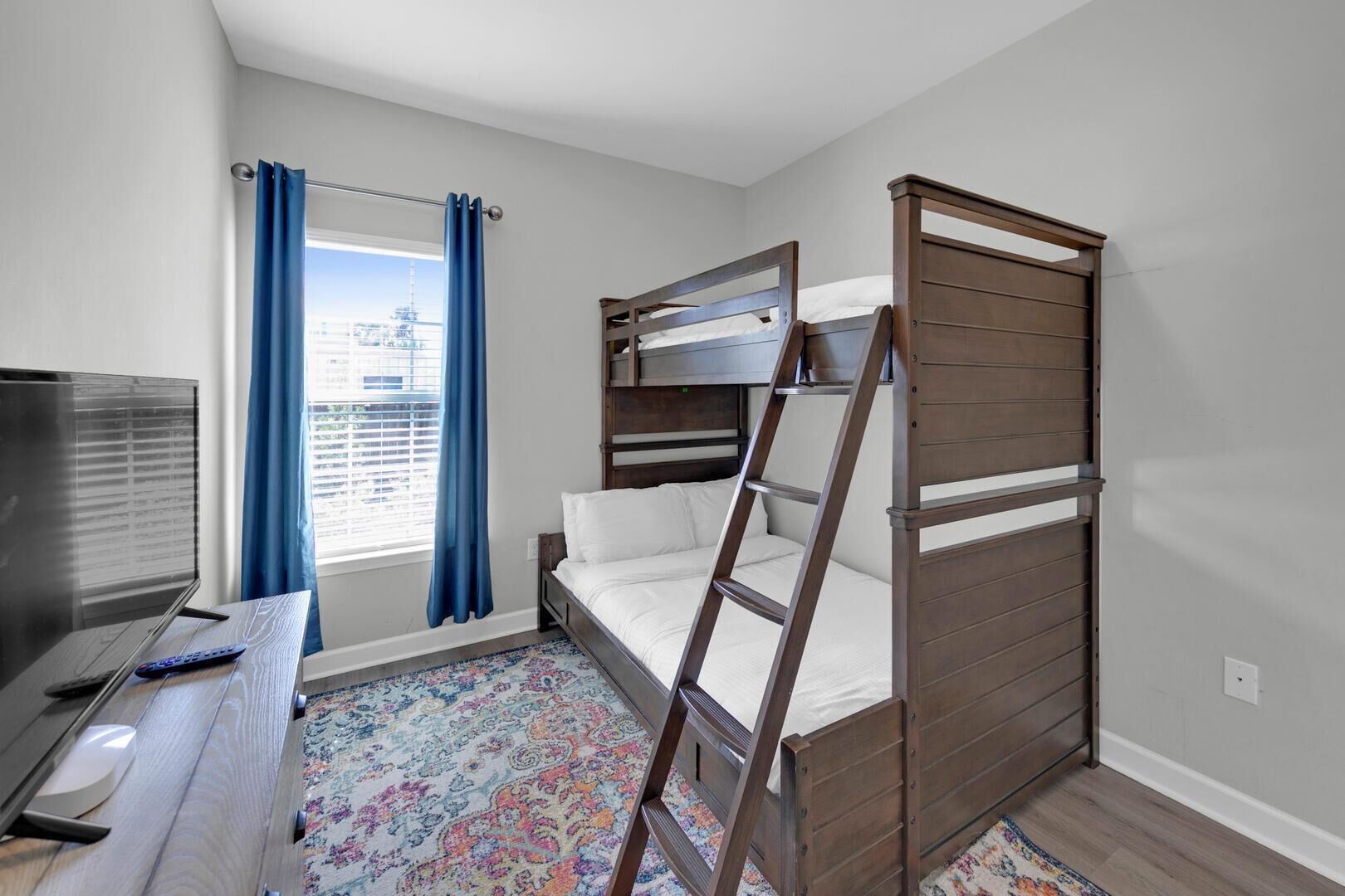 A compact room with a bunk bed, a desk with a television, and a window with curtains.
