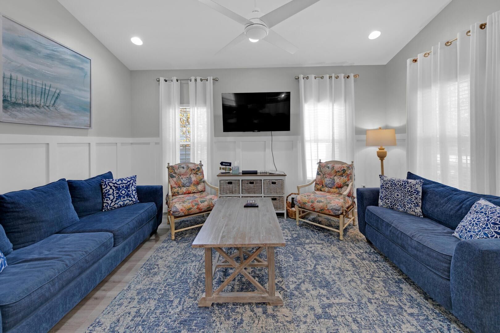 Ein Wohnzimmer mit zwei blauen Sofas, einem Holztisch und zwei floralen Sessel. Ein Flachbildfernseher ist an der Wand befestigt und auf einem Beistelltisch steht eine Lampe.