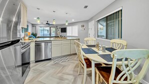 A modern kitchen with stainless steel appliances, a dining area with wooden chairs, and a large window providing natural light. - Crystal Village II, Unit 6B (Miramar Beach)