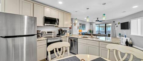 A modern kitchen with stainless steel appliances, a dining area with a round table and chairs, and a view of the living area through a glass door.