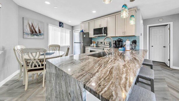 A modern kitchen with a marble island, white cabinets, and a dining area with wooden chairs. - Crystal Village II, Unit 6B (Miramar Beach)