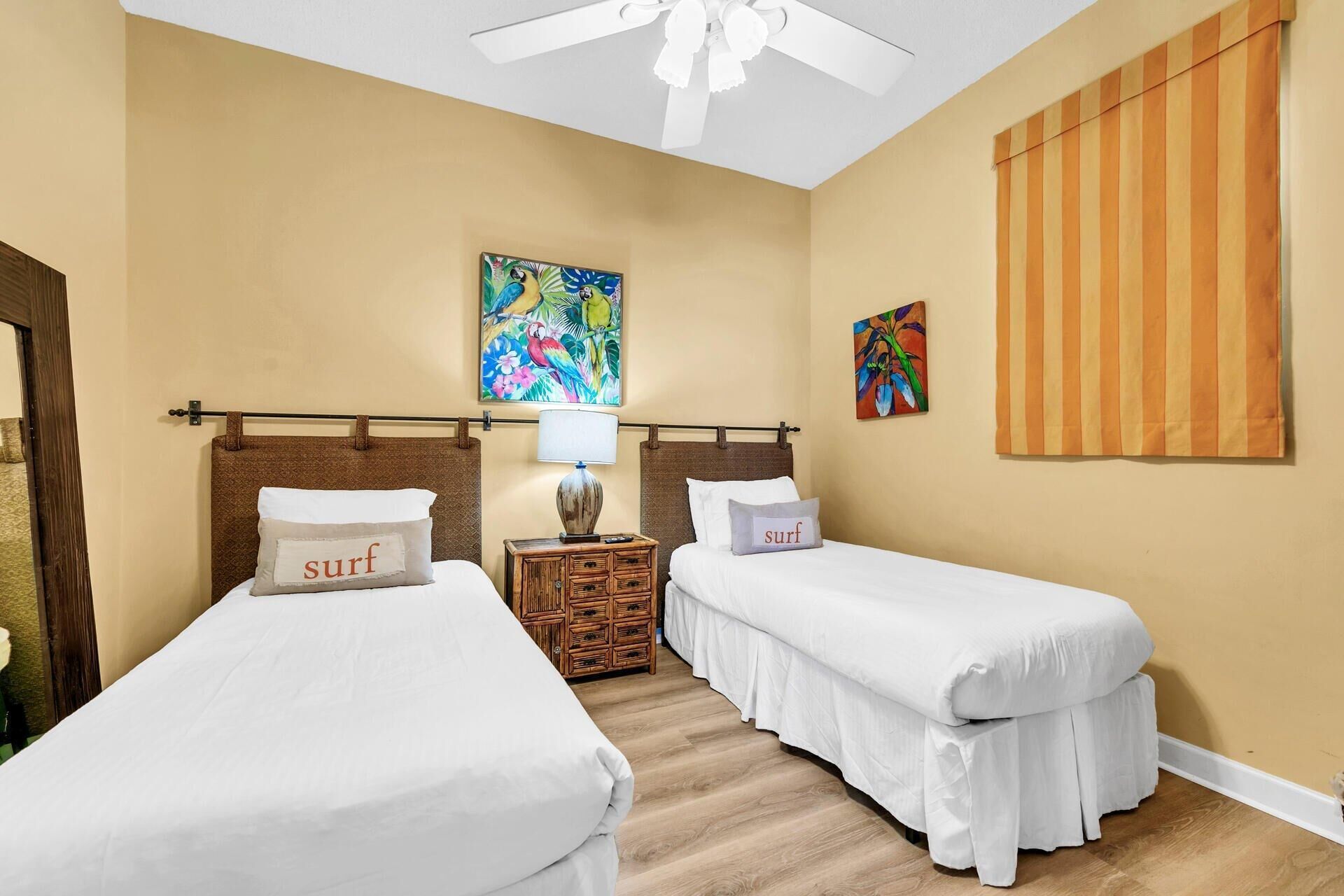 A hotel room with two single beds, a ceiling fan, a wooden nightstand with a lamp, and two colorful paintings on the walls.