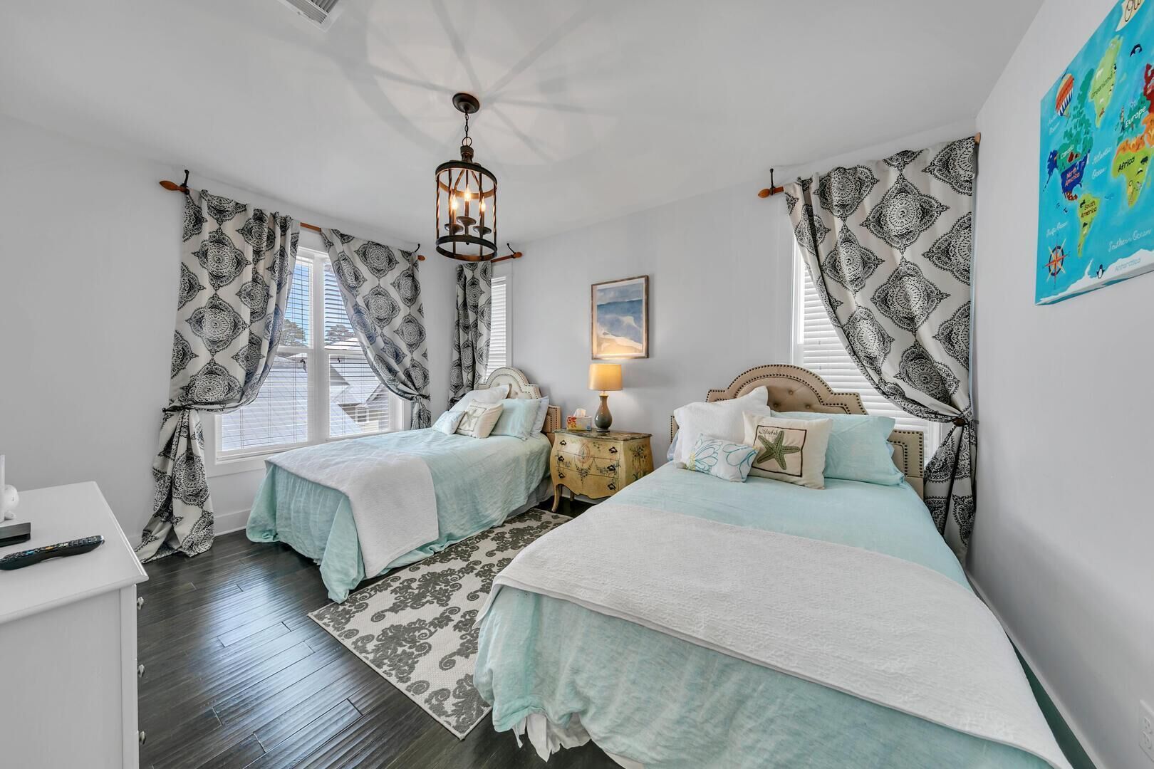 A bedroom with two beds, patterned curtains, a chandelier, a nightstand, and a map on the wall.
