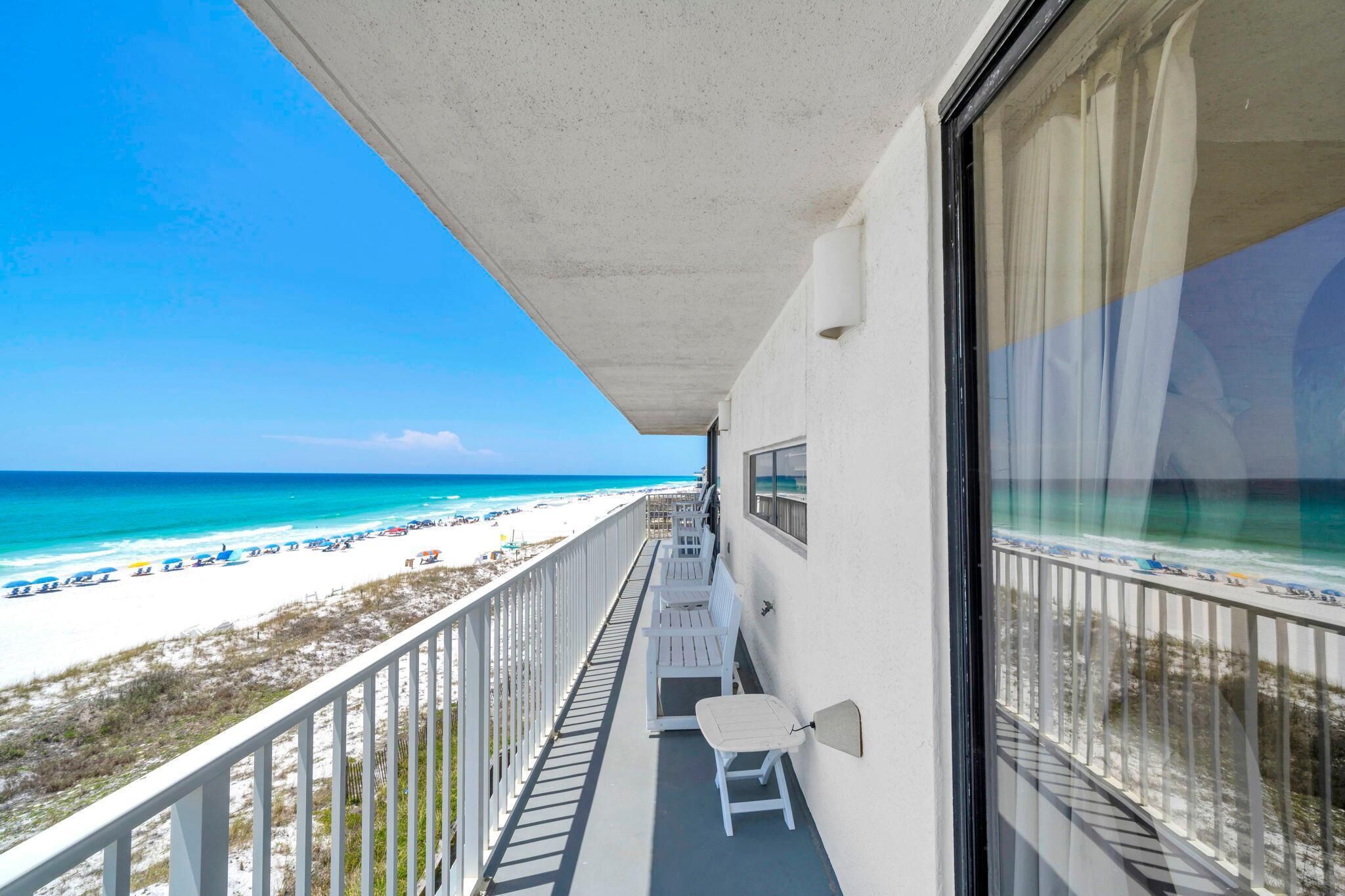 A balcony with a view of a beach, white chairs, and a railing.