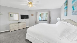 A bedroom with a large bed, a ceiling fan, a dresser, a TV, and a window with curtains. - Crystal Village II, Unit 18A (Miramar Beach)