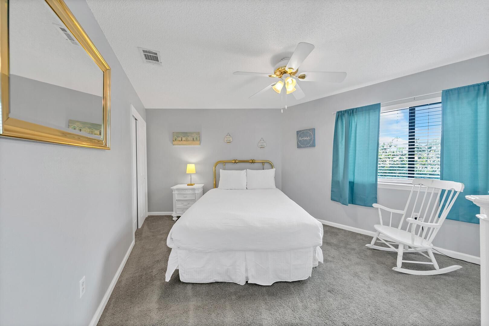 A bedroom with a bed, a nightstand, a lamp, a ceiling fan, a rocking chair, and a window with curtains.