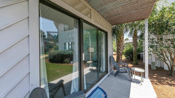 A covered porch with two chairs and a small table, a glass door leading to a patio, and a welcome mat with a palm tree design. - Crystal Village II, Unit 18A (Miramar Beach)
