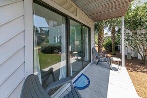 Un porche abrité avec deux chaises et une petite table, une porte vitrée donnant sur un patio, et un paillasson orné d’un motif de palmier.