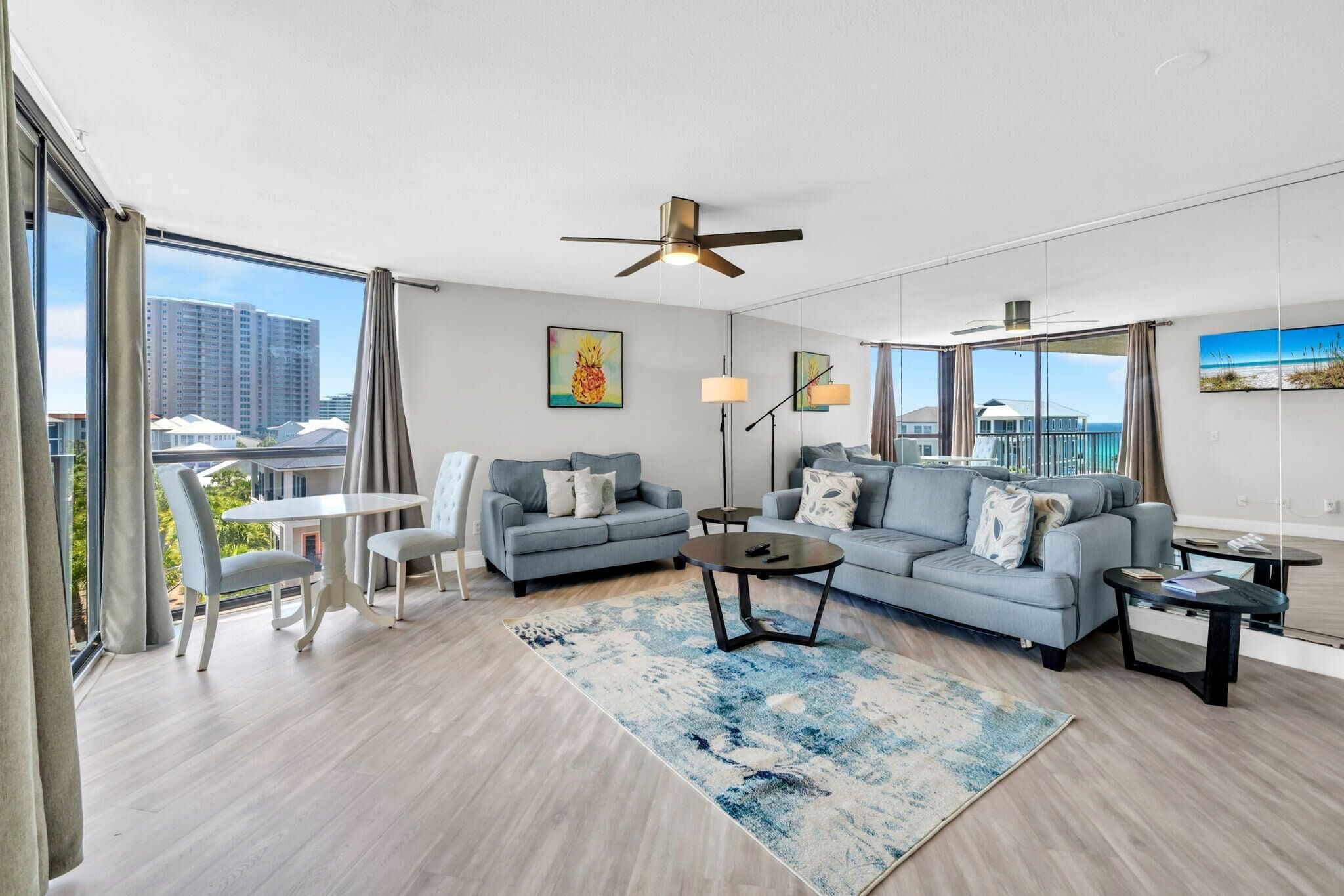 A modern living room with a sofa, coffee table, and dining area, large windows with curtains, and a ceiling fan.